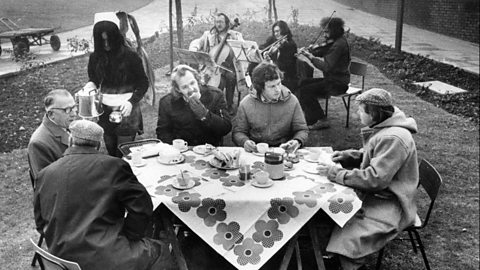 Outdoor tea party in a park with a floral tablecloth, cups and plates on the table, and a small group of musicians playing string instruments in the background.