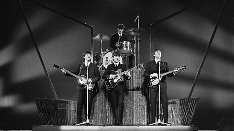 Iconic live performance of The Beatles on stage with guitars and drums, featuring their classic setup and instruments under dramatic stage lighting.
