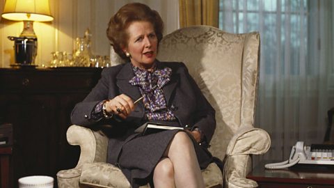 Person seated in an ornate armchair holding a pen and notebook, dressed in a formal suit with a patterned blouse, in a traditional living room setting with a lamp, telephone, and tea cup on a side table.