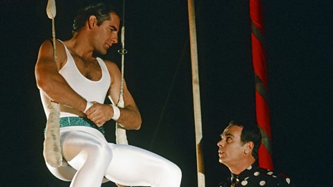 A still from Quantum Leap. Sam is dressed in all white and sat on a trapeze, looking down on Al
