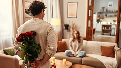 A man holding a bouquet of red roses behind his back for his partner who is sitting on the sofa in a cosy living room.