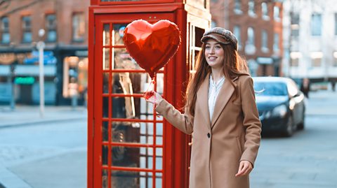 A young woman stands beside a bright red telephone booth, smiling as she holds a shiny heart-shaped balloon