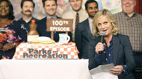 Amy Poehler as Leslie Knope in Parks and Recreation, next to a cake commemorating the 100th episode