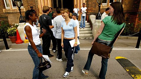 Group of students standing and chatting outside a brick school building, holding papers and bags, with others gathered near the entrance steps in a casual setting.