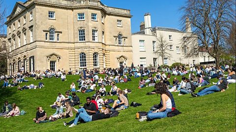Large group of students relaxing and studying on a grassy lawn outside historic university buildings under a clear blue sky on a sunny day.