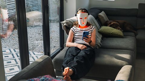 Person relaxing on a grey sectional sofa indoors, holding a smartphone and wearing headphones, with a dog lying on a cushion nearby and large glass doors letting in natural light.