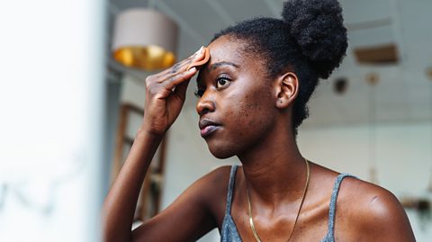 Person wearing a grey tank top and gold necklace indoors, with one hand raised to the forehead, and a modern pendant light in the background.