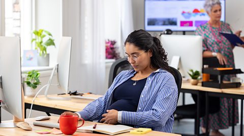 A pregnant woman working at her computer in an office. She has a hand on her stomach.
