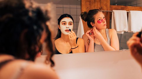 Two people standing in front of a bathroom mirror, adjusting earrings and getting ready, with towels hanging on a rack in the background