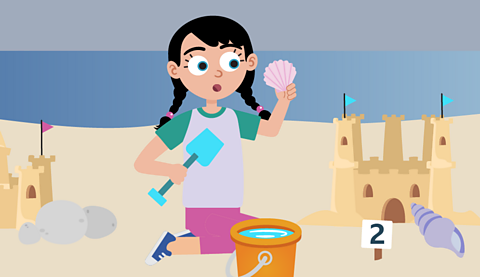 A girl looking panicked holding a shell and a spade in front of a sandcastle. She is on a beach and the sky is grey.