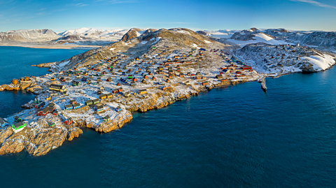 An aerial view of Inuit village in Greenland. The island is covered in some ice and surrounded by water. On the land are many colourful houses dotted around the village