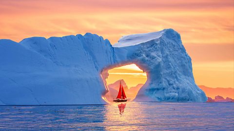 Midnight sun in Greenland. A boat with red flags travels on the water in the distance, making its way under an ice tunnel as the night sky is orange