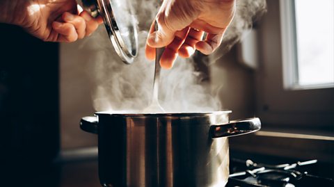 A silver pan is on a hob with steam pouring out. Inside the pan is soup. Image shows hands above the pan, the right holding a silver spoon and stirring and the left holding a glass lid which has been removed