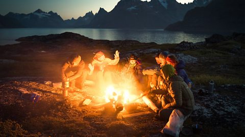 People talking around a campfire in Greenland. It is dark as the group of seven men sit on land in a semi circle around the fire, all wearing thermal such as big coats and hats. behind them you can see mountains in the distance