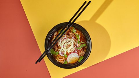 Bowl of broth wit h noodles and green herbs and a slice of radish on top. Chopsticks are on top of the bowl