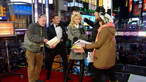  Ryan Seacrest, second left wearing a black suit and bow tie, next to Miss America Savvy Shields, blonde wearing a tiara and grey duffle coat, and staff either side with headphones ondo The Mannequin Challenge at Dick Clark's New Year's Rockin' Eve. They are on a stand in Time Square, New York, with huge buildings with advertising boards and crowds around them. They pose as if they are adding final touches to Savvy before they go live on air