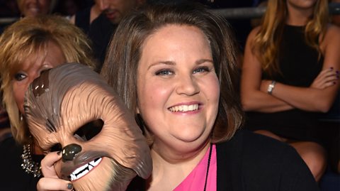Candace Payne, AKA Chewbaca Mom, holding her famous mask. She has short brunette hair and wears a pink dress and black cardigan. She smiles holding the mask in her right hand of the Star Wars character, who is a brown and neutral hairs beast with sharp teeth