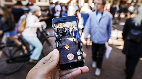 A crowds going about their day to day life on a street are blurred as the focus of the image is a smartphone. Through the screen is a Pokémon character on the street in non-blurred video, as the person plays on the Pokémon Go app