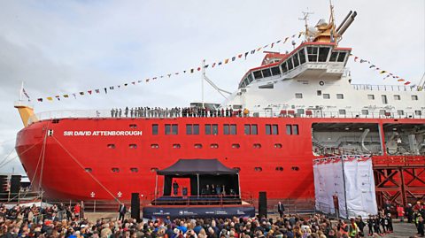 Crowds gather around the docked RRS Sir David Attenborough. The huge ship is red with white text which reads " Sir David Attenborough." The top half of the boat is white and over a dozen people stand on the top deck overlooking the crowd.