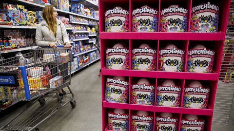 Hatchimal toys, animals which hatch out of an egg, on a pink stand on display inside an American supermarket. To the left is another shop aisle filled with toys on the shelves. A woman pushing a trolly stands in the aisle browsing what's on offer