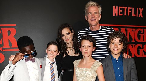 Stranger Things first aired back in 2016. Pictured, actors Caleb McLaughlin - wearing sunglasses and a grey and white suit, Noah Schnapp - in a grey suit and tie, Winona Ryder in a black dress with long black hair,, Millie Bobby Brown - wearing a cream patterned dress with short brown hair, Matthew Modine in a white and black striped long-sleeved top and and Gaten Matarazzo, in a grey suit jacket and navy blue shirt, attend the premiere of Stranger Things. Behind them is a black promotion board for photos, with the bold red Netflix Stranger Things logo 