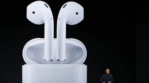 Apple Senior Vice President of Worldwide Marketing Phil Schiller announces AirPods during a launch event. He has blond hair and wears a navy blue shirt on stage while presenting. Behind him is a huge screen showing with white Air Pods, two white earphones coming out of a small pod, against a black background