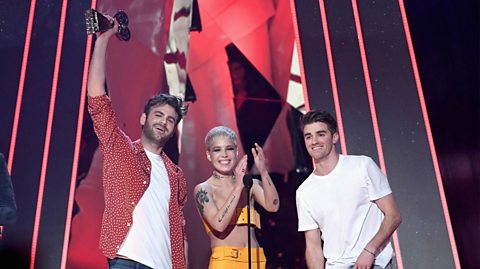 Artists Chainsmokers and Halsey who released 2016 hit Closer. Holding an award on stage is DJ Alex Pall, wearing a red shirt and white top, next to singer Halsey, who has short blonde hair and wears a yellow two piece outfit while applauding and DJ Drew Taggart, far right in a white top
