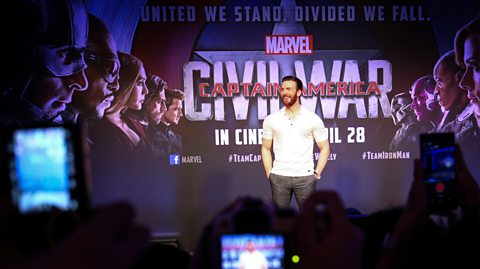 Chris Evans at the Captain America press launch, 2016. He has slicked back brown hair and a beard and smiles wearing a white short sleeved top and grey washed out jeans. Behind him is a promotional poster for the film and out of focus in the forefront of the picture is a crowd, some holding camera phones and capturing the moment