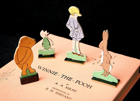 A pale orange Winnie-the-Pooh book with characters from the children's book standing on top of it. Left to right they are Winnie-the-Pooh, a yellow bear with his hands held behind his back, piglet - a small pale pink pig with a green striped top on, Christopher Robin - a young boy with blonde hair, a yellow hat and a pale blue jacket and shorts, and Kanga - a pale brown kangaroo