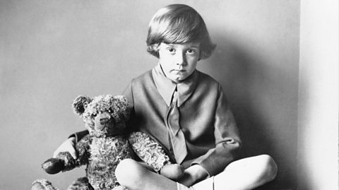 A black and white photo of Christopher Robin as a child, with long sweeping hair and wearing a scouts style uniform - a tie, shirt and shorts. He sits crossed-legged on the floor with a blank expression looking to the camera. On his left is his toy teddy bear