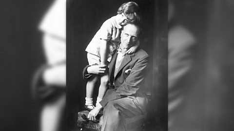 A black and white photo of A.A. Milne and his son, Christopher Robin. Milne wears a suit and dickie bow, sitting on a wooden stool smiling at the camera. Standing on the stool to his left is his son, who he wraps his arm around his legs and hold his hands. Christopher has long hair and is wearing a short sleeved shirt, shorts and plain shoes with socks. He looks at the camera and rests his head on to his dad's, hugging him with his left arm