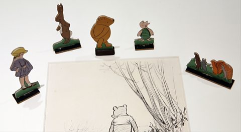 Original Winnie-the-Pooh illustrations. Image shows a piece of paper centre, with a black and white sketch of Pooh bear with his back to the viewer, walking in the forest. Around the paper are a number of illustrated characters stood up. Left to right, this includes Christopher Robin - wearing a yellow hat with blond hair and wearing a light blue jacket and shorts, Kanga - a light brown kangaroo, Pooh - a yellow bear resting his arms behind his back, Piglet - a small pale pink pig wearing a green jumper and other forest animals to the right, including a squirrel and hedgehog