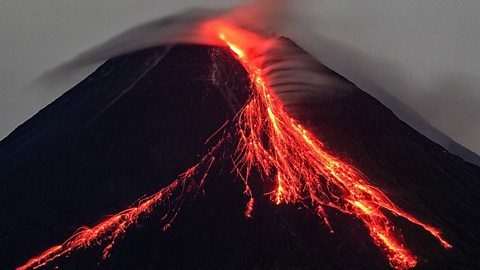 Active volcano erupting at night with bright red lava flowing down the slopes and ash clouds rising from the crater.