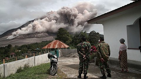 Group of people watching a volcano release a massive ash plume over nearby homes and vegetation.