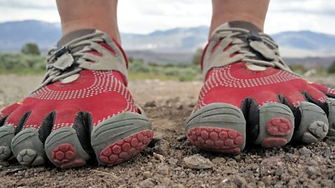 Close-up view of a person wearing red and grey five-toe minimalist running shoes while standing on a rocky outdoor trail, with blurred mountains in the background.