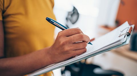 A person holding a clipboard with a pen wearing a yellow mustard shirt in a blurred office background