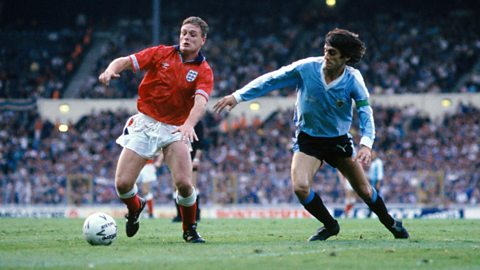 Paul Gascoigne, wearing the 1990 red Umbro away kit, dribbles away from a Uruguayan defender in a friendly at Wembley