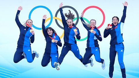The 2022 British women's curling team jump in the air holding their Winter Olympic gold medals with the Olympic rings behind them