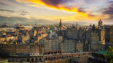 An aerial view of Edinburgh city centre, showing tonnes of old buildings and a huge clock tower in the distance. The sky is bright with a yellow haze