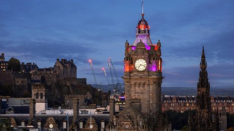 The sky darker as evening approaches in Edinburgh city centre. An aerial view shows the top of a number of old buildings but the focus of the image is the centre, the Balmoral Hotel clock tower. The dark brown clock tower is illuminated with purple and orange lights at the top