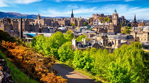 A view over historic Edinburgh from Carlton Hill. At the forefront of the image is a path lined with trees, one side the leaves bright green and the other side, autumnal orange and brown tones. Behind is the city full of old buildings, in the distance the Balmoral Hotel clock tower and Edinburgh Castle which sits on the hillside