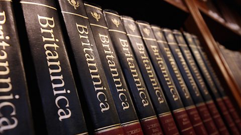 A dark wooden bookshelf is lined with thick Britannica encyclopaedias. The are all black with maroon colouring at the bottom declaring which volume. Along the spine of each book reads 'Britannica' in white bold text