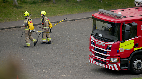 In Edinburgh, two members of the fire service wear their pale uniforms with illuminous stripes and helmets as they carry equipment to a scene. The leave behind them a red fire engine parked up on the side of the road