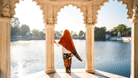 Getty Images Woman with orange headscarf stands between carved stone arches looking out onto a tranquil lake in Udaipur (Credit: Getty Images)