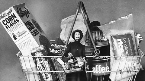 A black and white photos of a woman in the 1970s standing and holding a metal shopping basket. She has curly, short dark hair and wears and dark, high neck long-sleeved mini dress. In her basket are items like a Corn Flakes cereal box, a tin of Heinz Beans and a bag of long-grain rise. Behind her is an enlarged photo of the same basket with the same items she holds