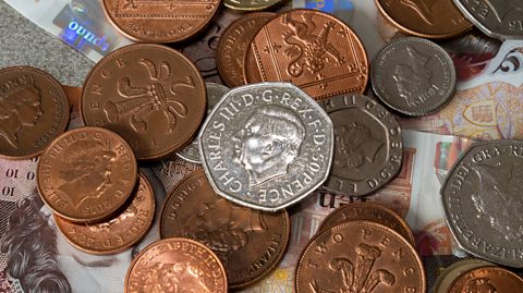 A handful of coins lie on top of notes on a table. The coins and notes are British Pound Sterling. The notes are £10 notes and are orange and white and shiny, with a portrait of Queen Elizabeth II from the neck up, wearing a crown and with curly hair. The coins are 1p and 2p in a bronze colour, as well as some silver 50ps, 5ps and 20p. In the centre is a shiny silver 50p with a portrait of King Charles III in the centre. The portrait is a side profile of the King's head, with short hair