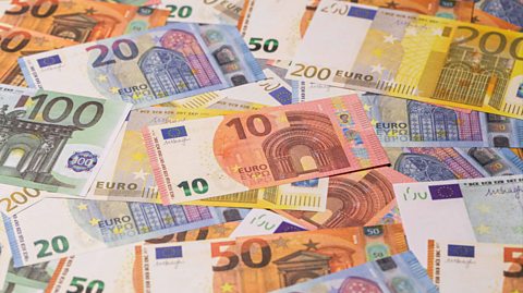Various euro notes lay on top of one another in a scattered pile. The 10 euro notes are pink and cream, the 20 notes are light blue and cream, the 50 notes are bright orange and cream, the 100 notes are light green and cream and the 200 euro notes are vibrant yellow and cream