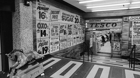  Tesco supermarket at The Bullring, Birmingham, in 1975. The black and white photo shows the entrance to the supermarket, with stairs and rails leading down into the store. Above the open door reads 'Tesco' and decorating the windows and walls on the outside of the shop and through the entrance are various items listed and their prices. Some of the prices include pre-decimalised coins, such as the half penny. At the forefront of the image is a children's rocking horse machine with a penny slot to activate