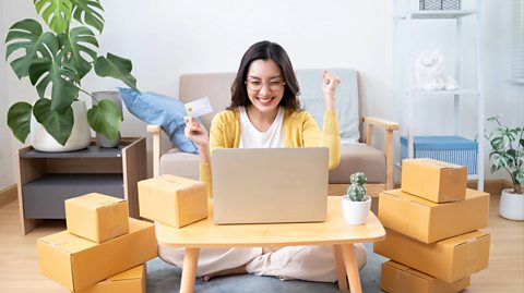 A young woman with long, black hair wearing glasses, a white top, a yellow cardigan and cream jogger pants sits crossed legged on the floor of a living room. Surrounding her is a wooden stand with a plant pot, a sofa behind her, a shelf stand and piled up light brown boxes. In front of her is a small coffee table with a small cactus and a silver laptop on the table. She smiles ecstatically at the screen, clenching her first with her left hand in the air and in her right hands holds a debit or credit card, after making an online purchase 