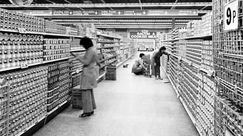 Inside a Co-op supermarket in Stoke, 1975. The black and white image shows one supermarket aisle, with shelves on both sides fully stocked and neatly stacked with items, mainly tins or food. Down the aisle are a number of customers scanning the shelves